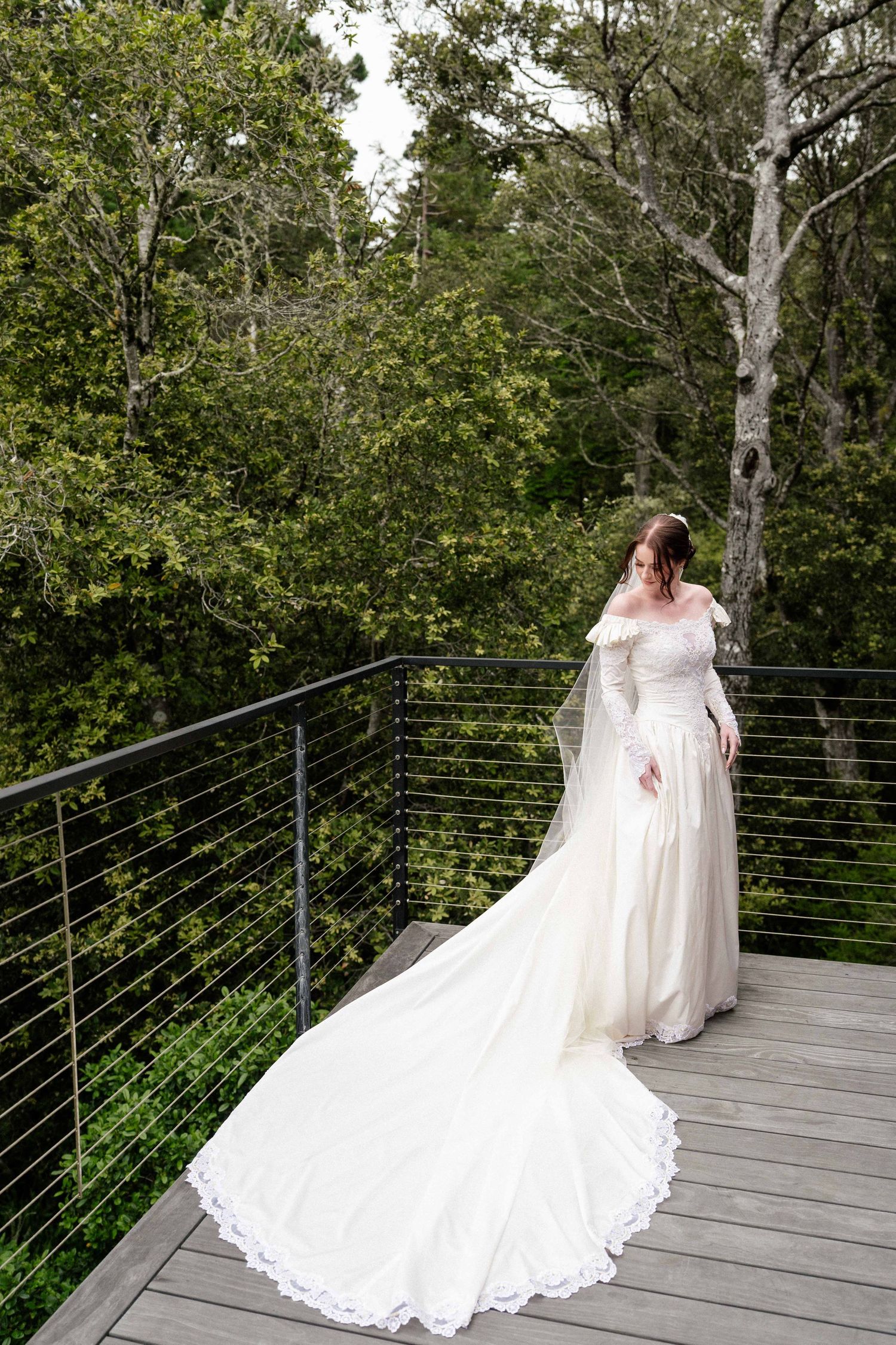 Bride in white off-shoulder gown with long train and veil stands on wooden deck surrounded by lush green trees.