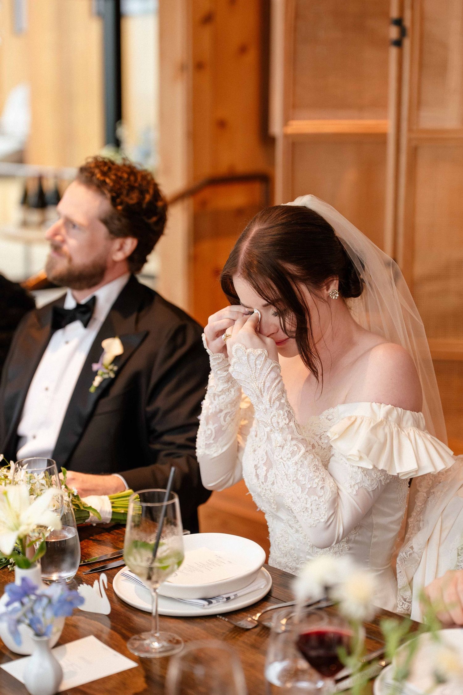 Emotional bride in lace off-shoulder gown crying at wedding reception table beside groom in black tuxedo.