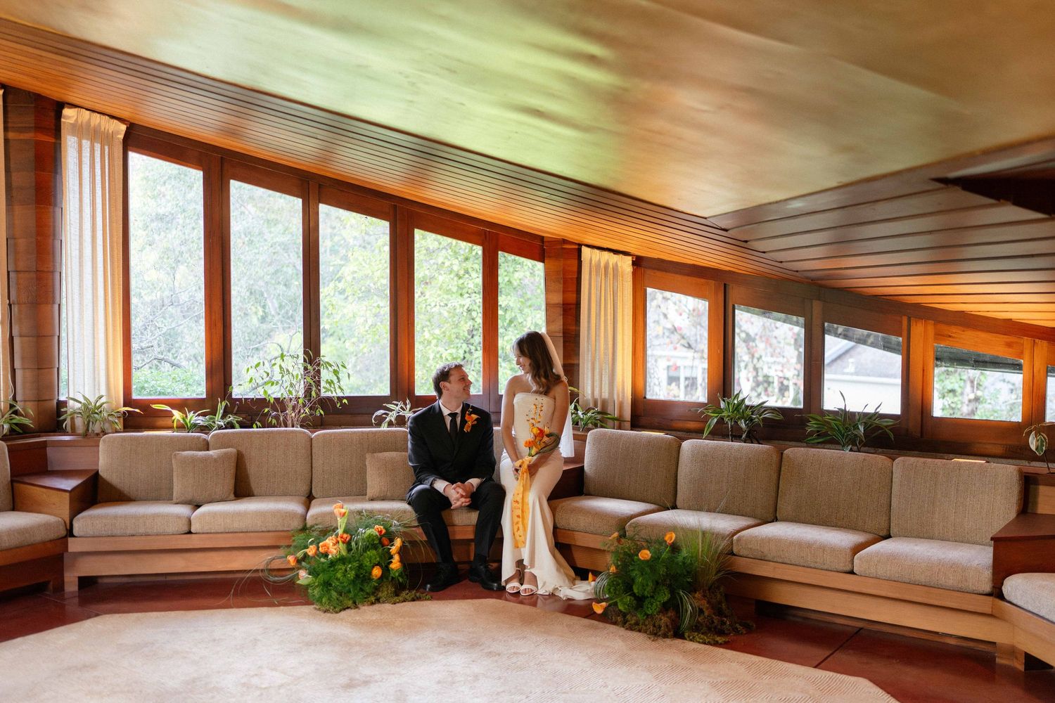 A bride and groom share a quiet moment on a mid-century modern sofa in a wood-paneled room with garden views.