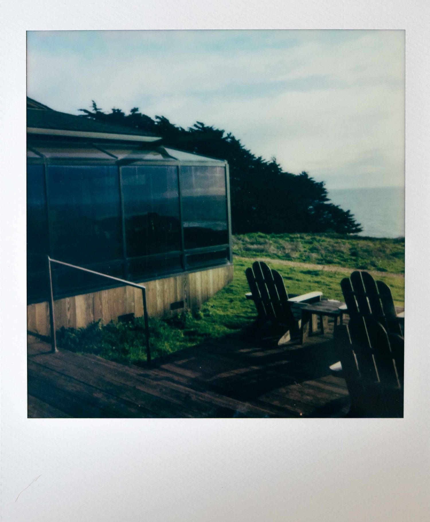 Two Adirondack chairs on a wooden deck overlooking the coast with a glass-enclosed structure nearby.