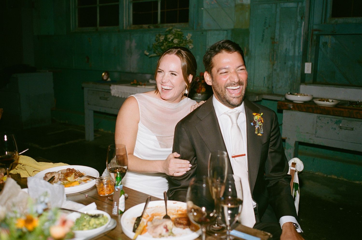 Bride and groom laugh joyfully together at wedding reception dinner table with food and drinks.