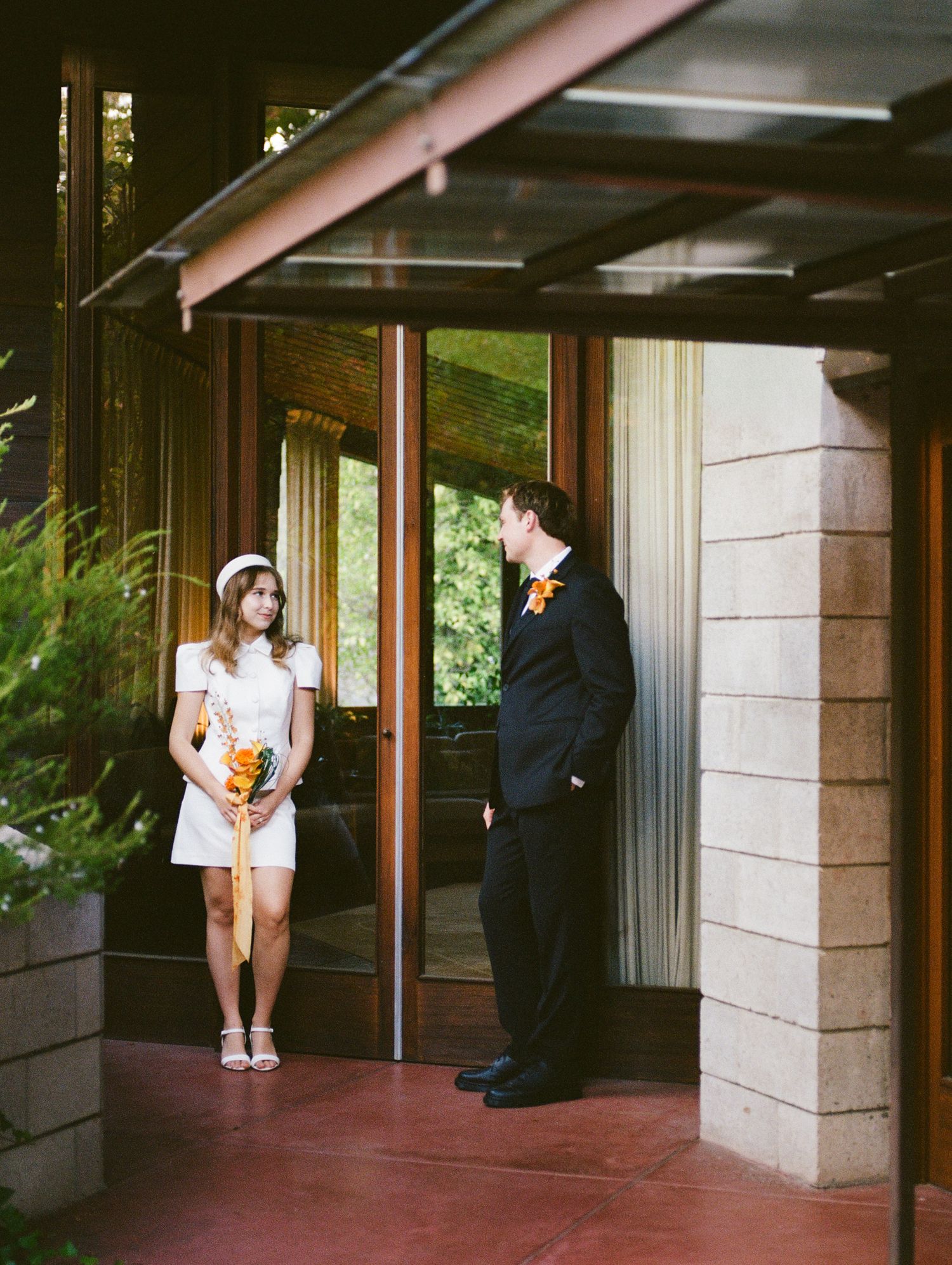 Bride in short white dress and groom in black suit sharing a moment at a mid-century modern building entrance.