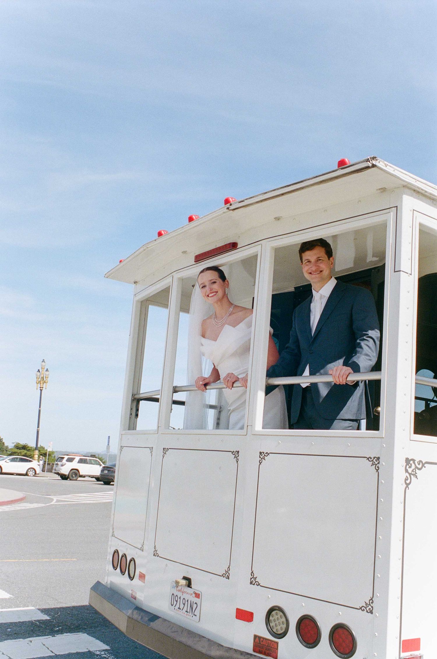 Bride and groom smiling on a white vintage trolley under a blue sky on their wedding day.