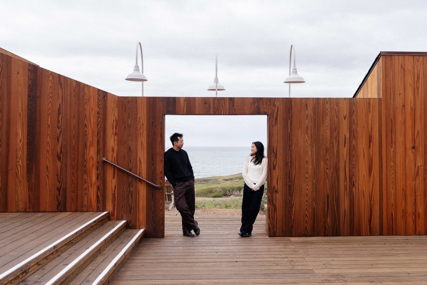 Two people stand in a timber-framed outdoor doorway overlooking the ocean on an overcast day.