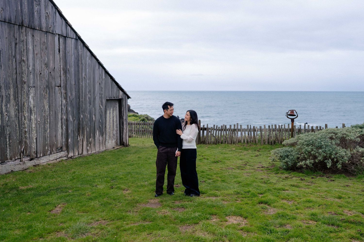 Couple standing on green grass beside a rustic wooden barn with ocean views under overcast skies.