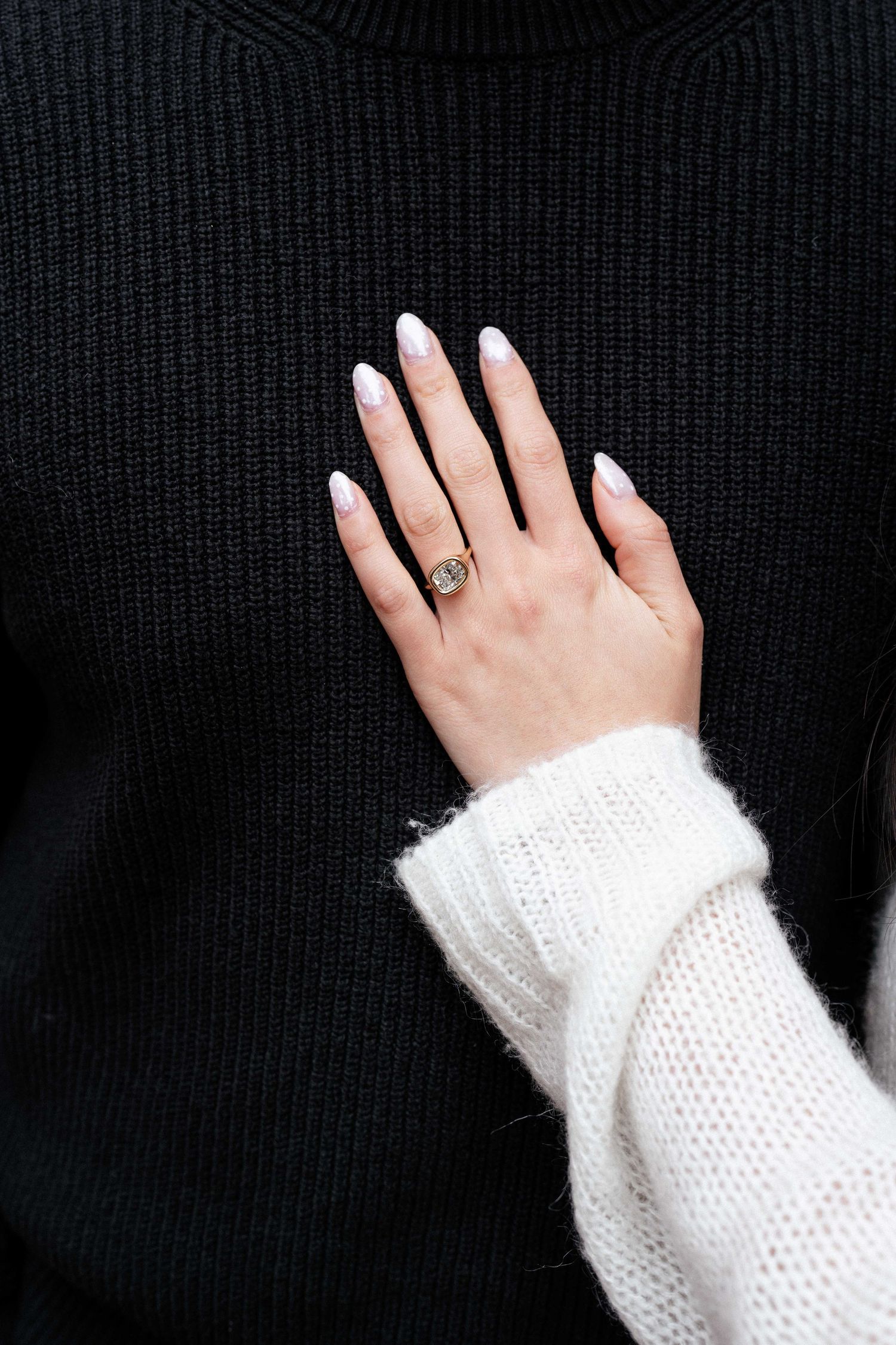 A hand with white French tip nails wearing a gold signet ring, resting on a black knit sweater, with a white fuzzy sleeve.