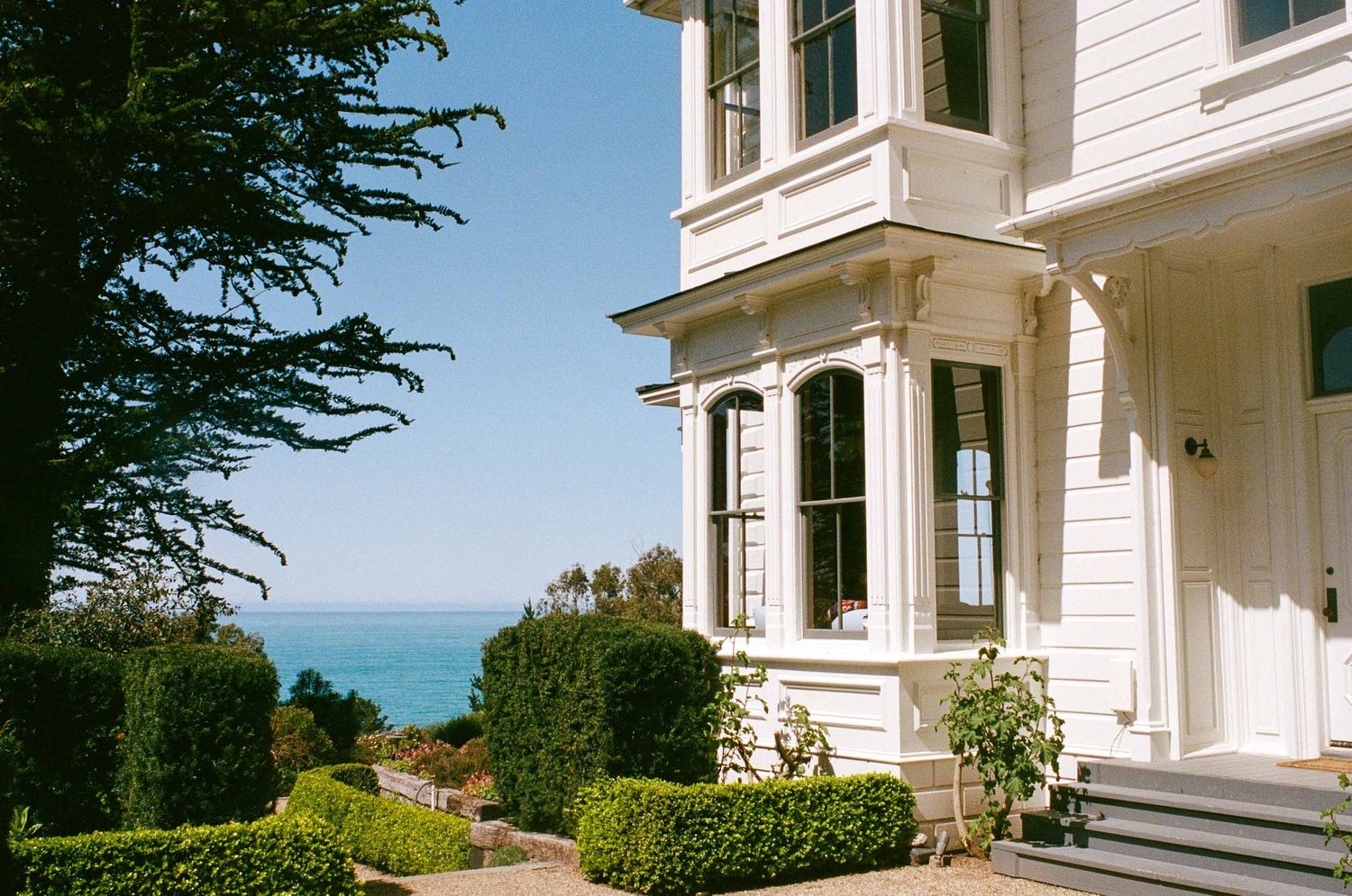 Victorian-style white coastal home with bay windows and manicured hedges overlooks the blue ocean on a sunny day.