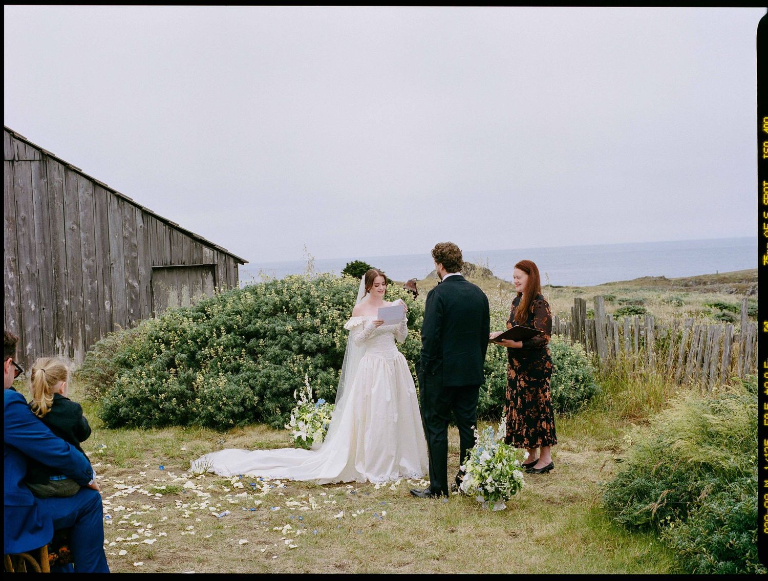 Outdoor coastal wedding ceremony with bride in white gown, groom in suit, and officiant by a rustic barn.