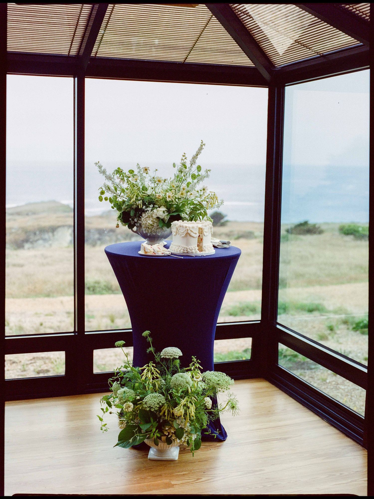 Elegant blue cocktail table with floral arrangements in a glass conservatory overlooking coastal scenery.