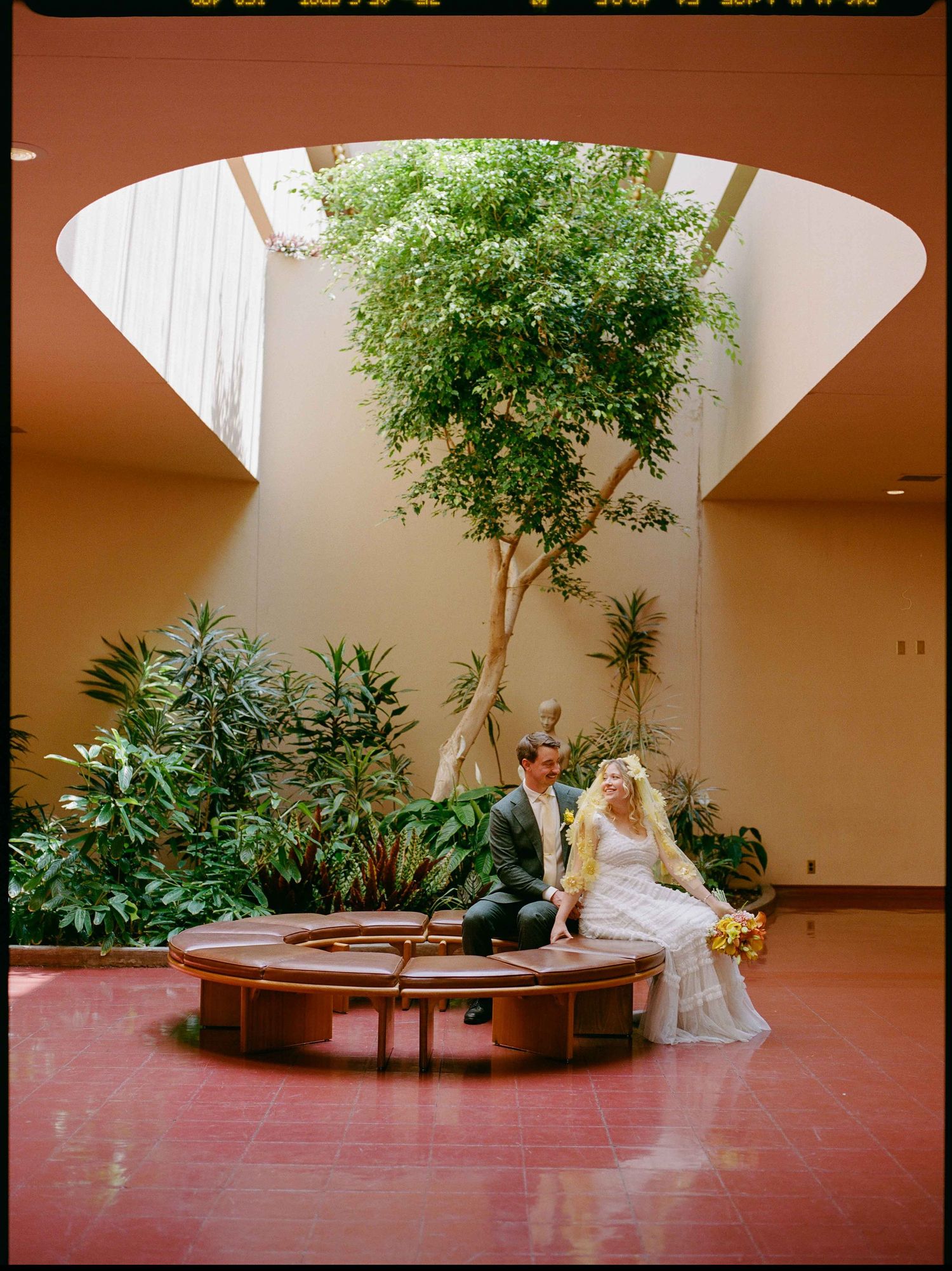 Bride and groom sitting on a circular bench in an atrium with lush greenery and a large skylight above.
