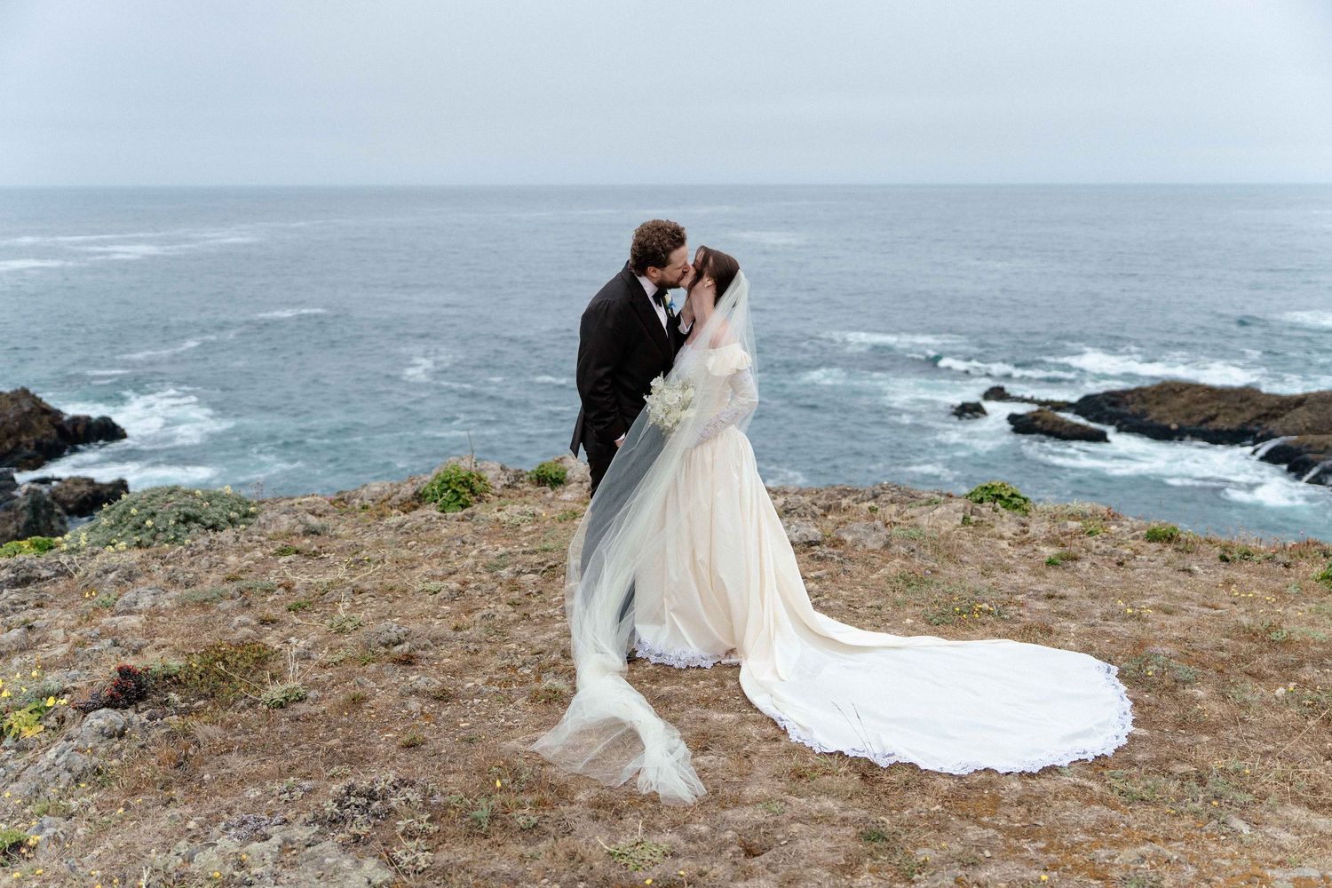 Bride and groom share a romantic kiss on a coastal cliff overlooking the ocean, with her white gown and veil flowing in the breeze.