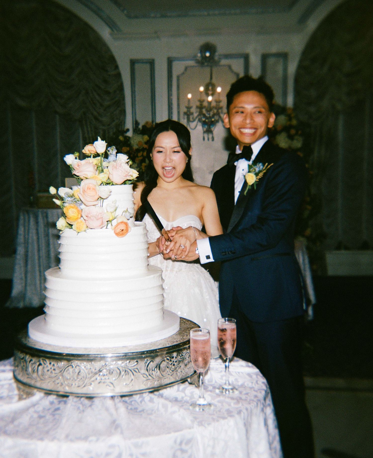 Smiling newlyweds cutting a white three-tier wedding cake adorned with pastel flowers, champagne flutes nearby.