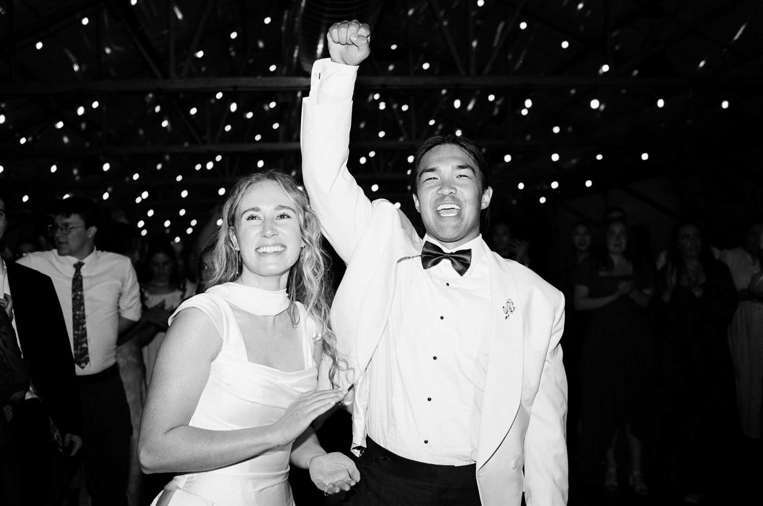 Black and white photo of a joyful bride and groom dancing at their wedding reception, groom raising fist in celebration.