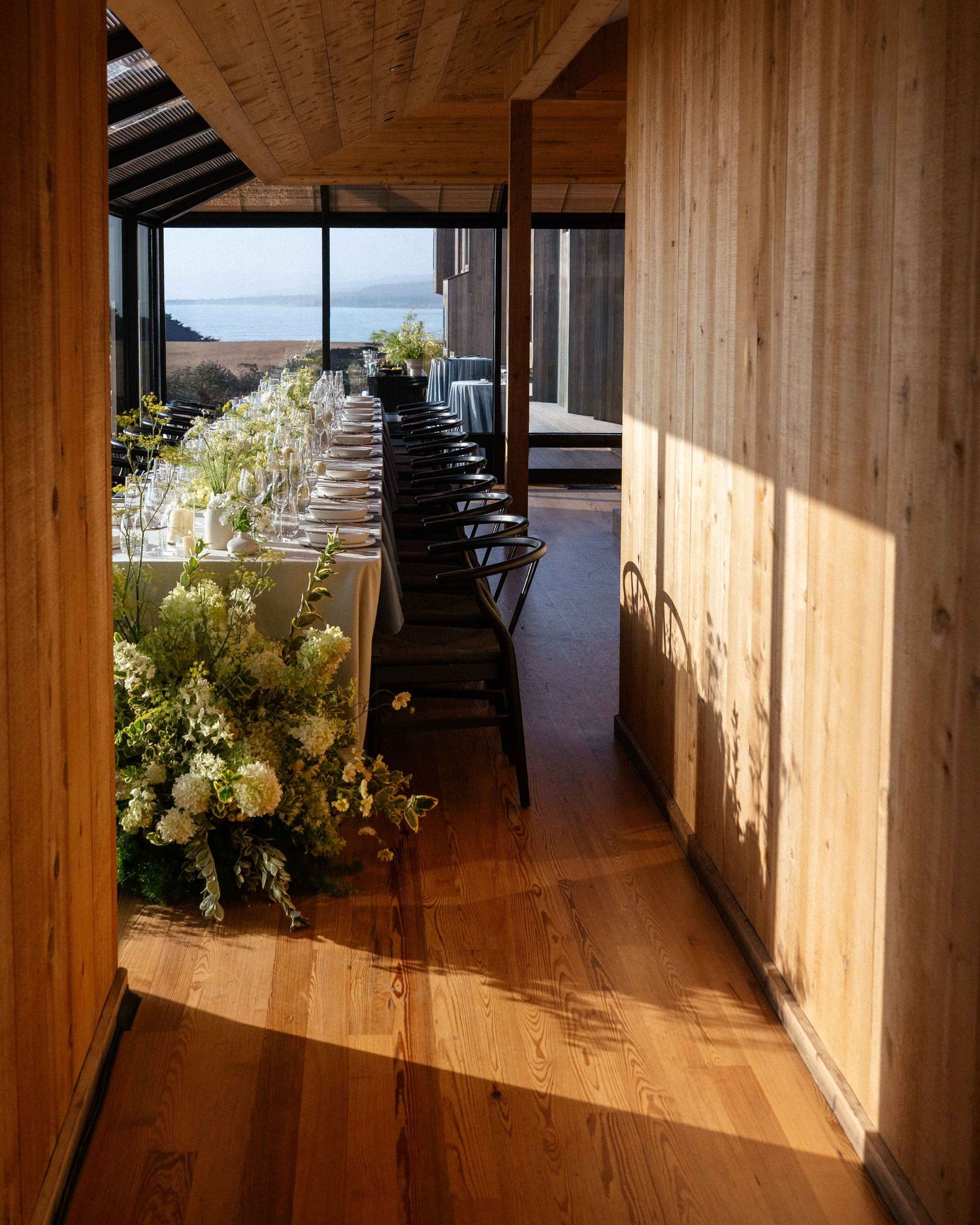 Elegant wedding reception with long floral table, wood-paneled walls, black chairs, and ocean views in warm sunlight.
