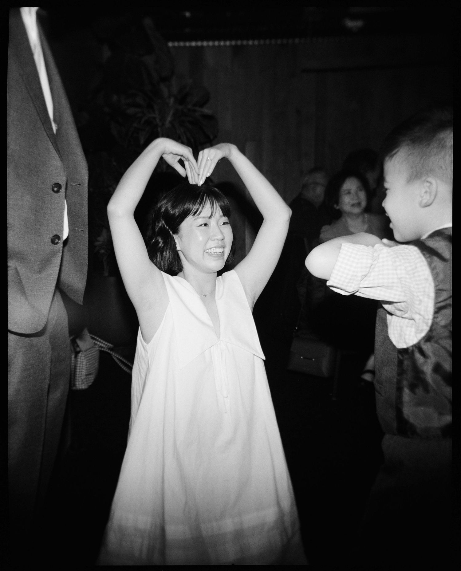 A young girl in a white dress makes a heart shape with her hands overhead while dancing at a party.