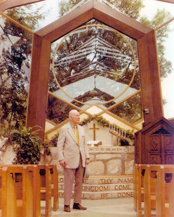 Man standing in a unique hexagonal glass chapel entrance with wooden beams and a cross inside.