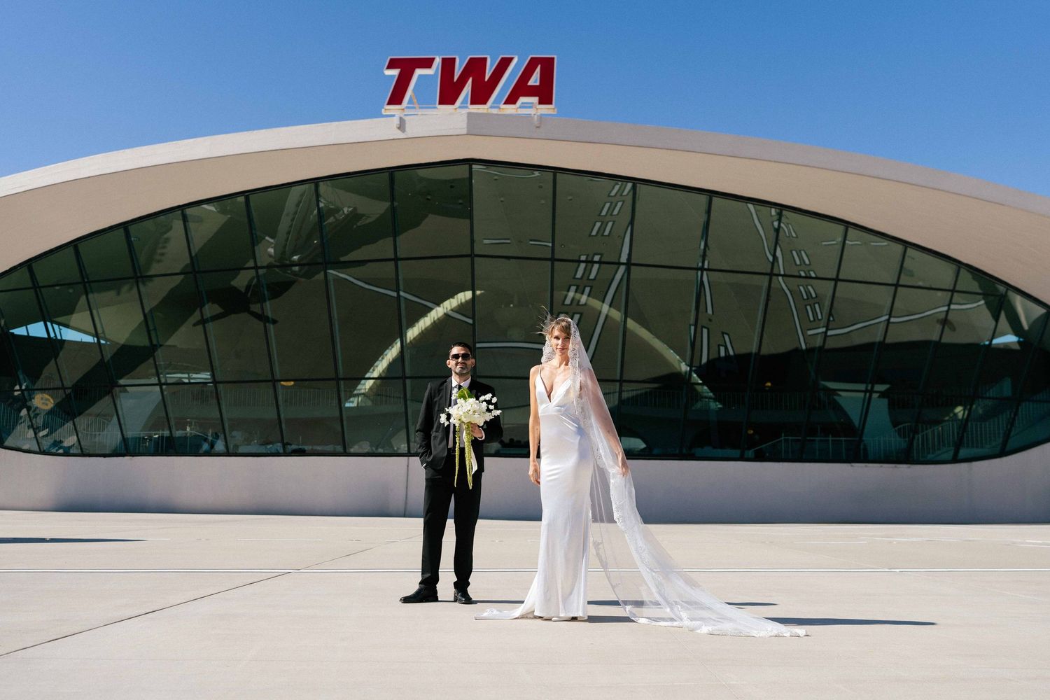A bride and groom stand in front of the iconic TWA Hotel terminal at JFK Airport on their wedding day.