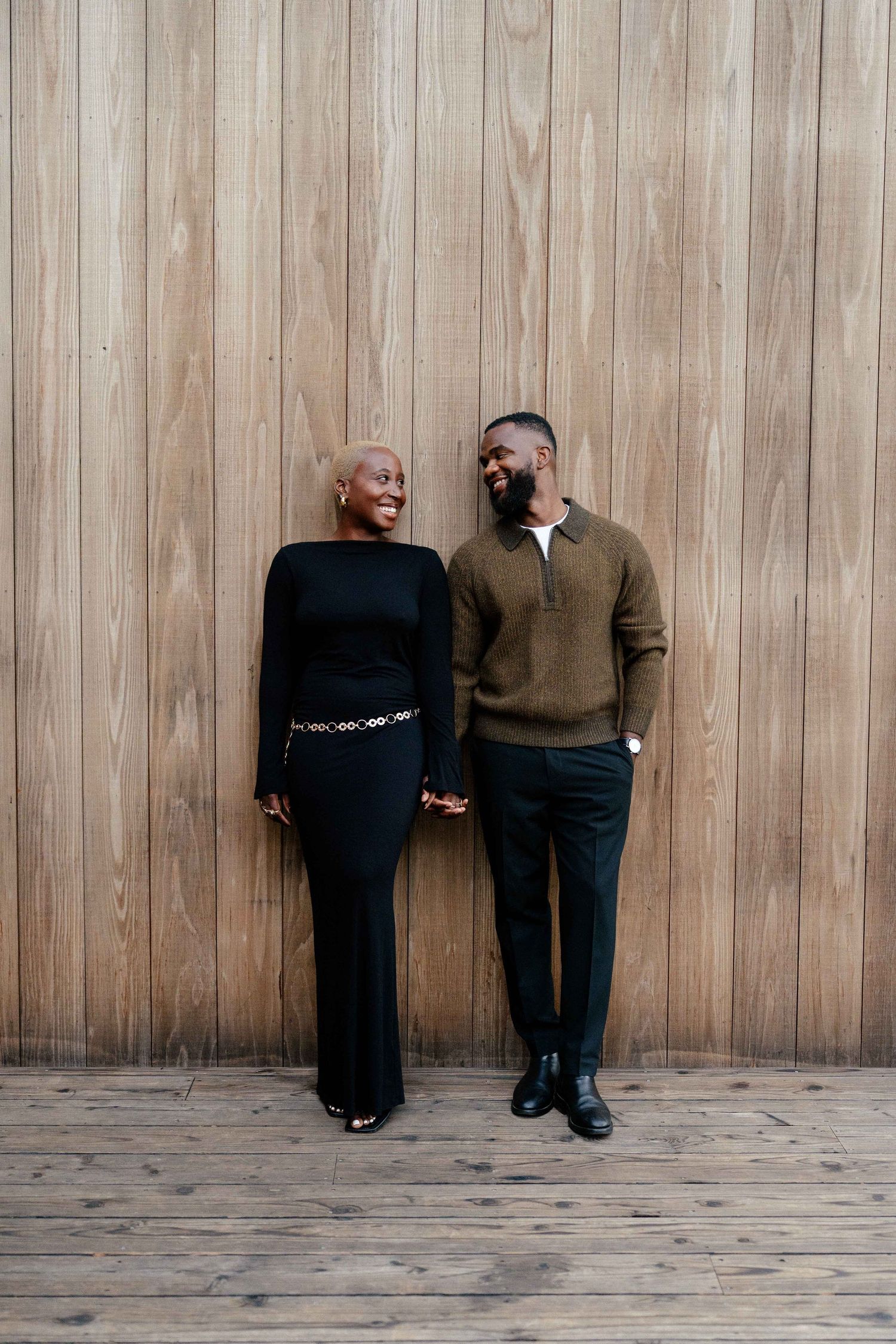 A joyful Black couple holds hands and smiles at each other against a warm wooden wall backdrop.