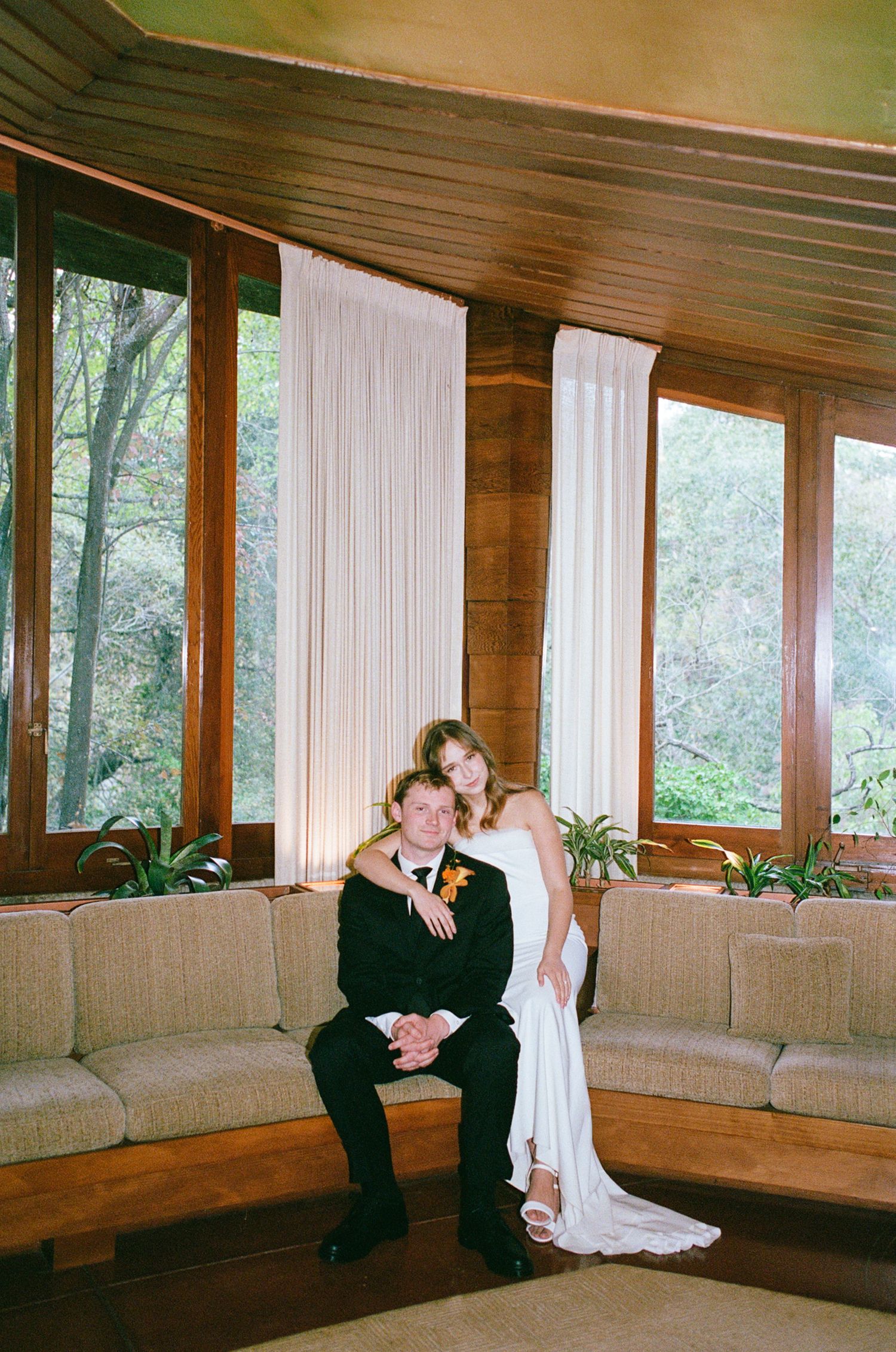 Bride and groom pose together on a couch in a mid-century modern room with large windows and lush greenery.