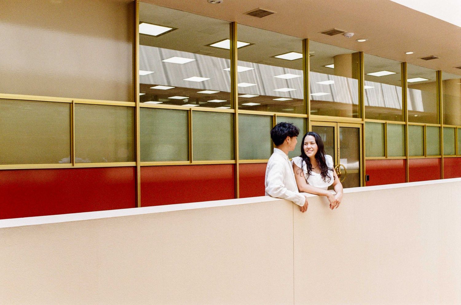 Couple sharing a romantic moment on a balcony overlooking large windows with gold frames in a modern building.