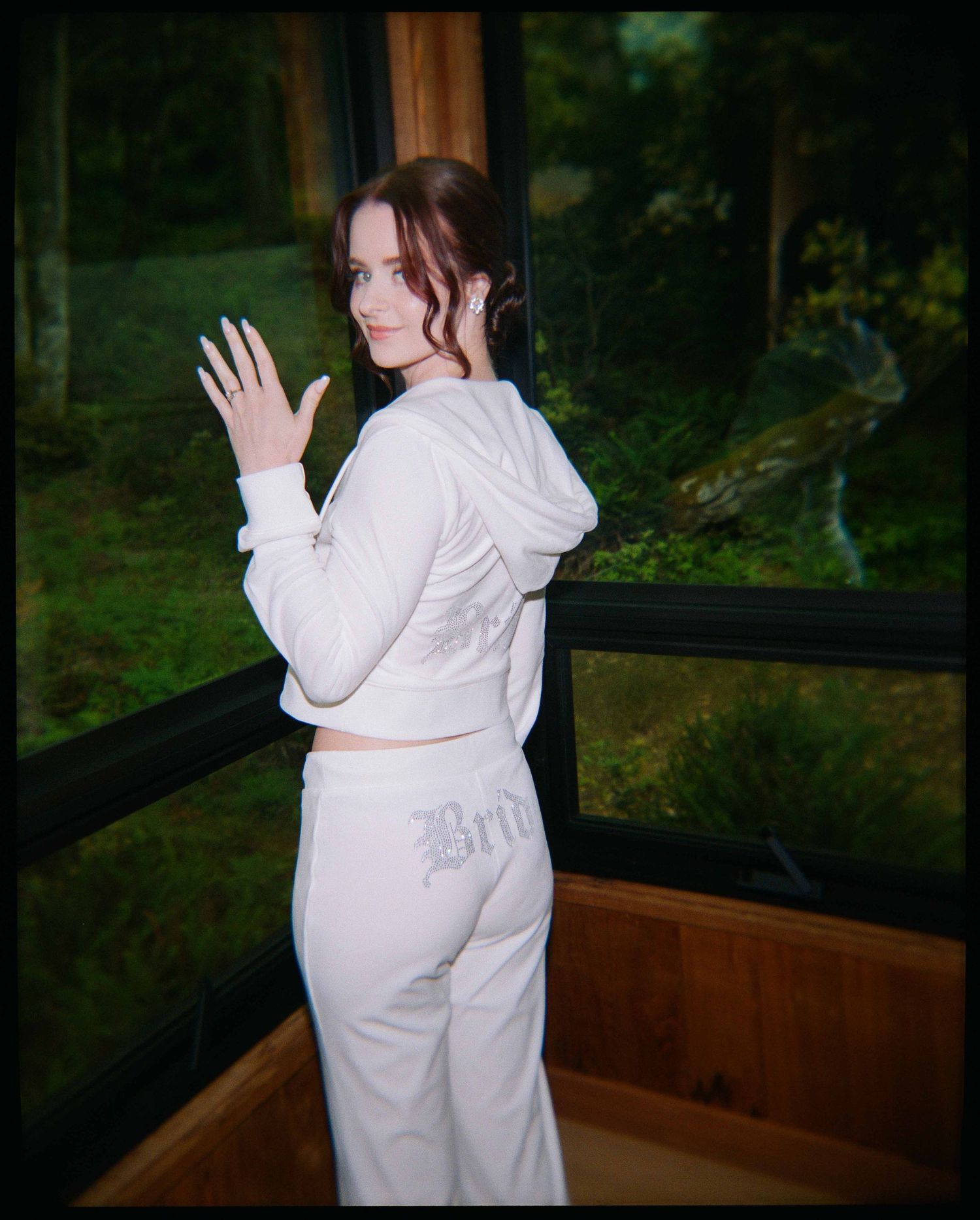 Young woman in white velour tracksuit posing indoors near large windows with forest view.