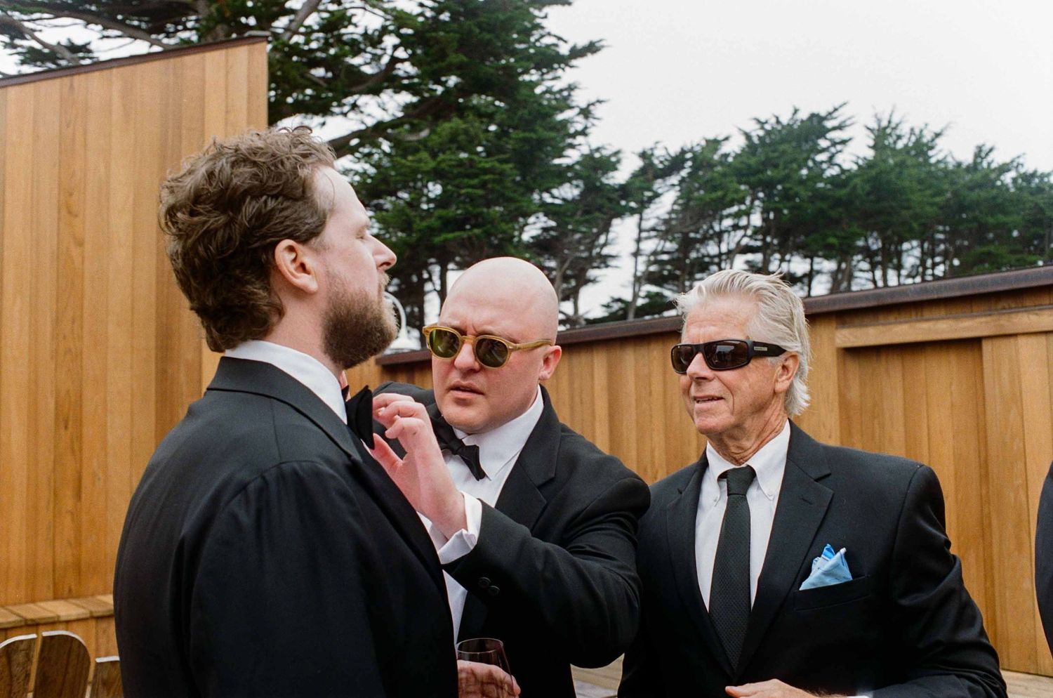 Three men in black suits adjusting ties outdoors at a wedding, with trees and wooden fence in background.