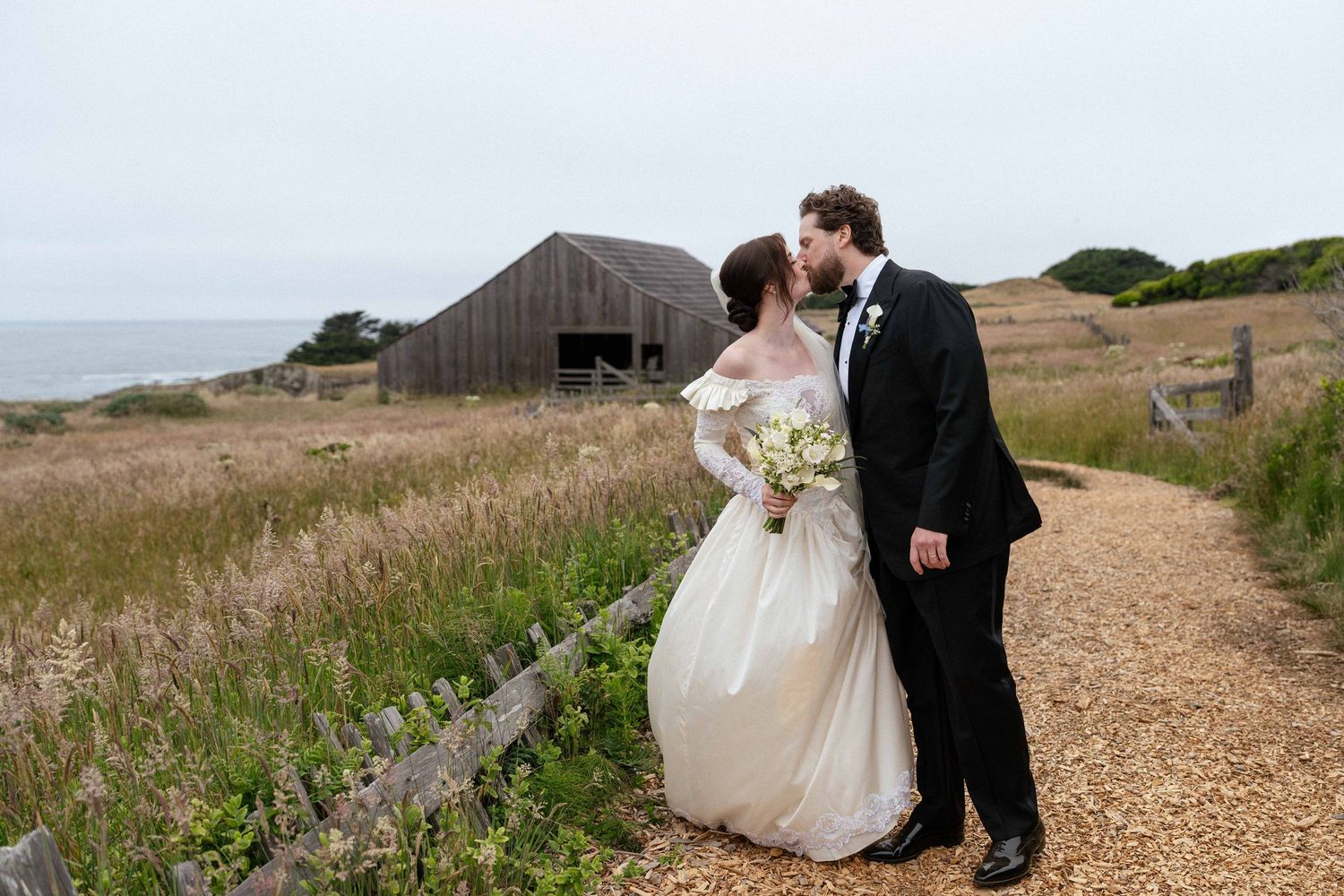 Bride and groom kissing on a scenic coastal path with rustic barn and ocean views on their wedding day.