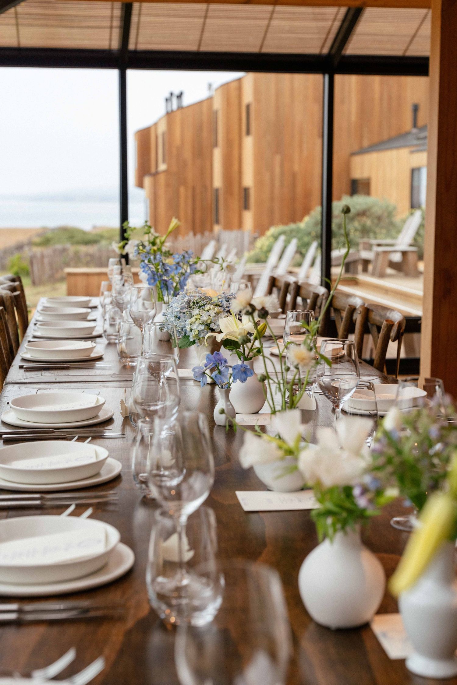 Elegant long dining table set with white plates, glassware, and blue and white floral arrangements near large windows.