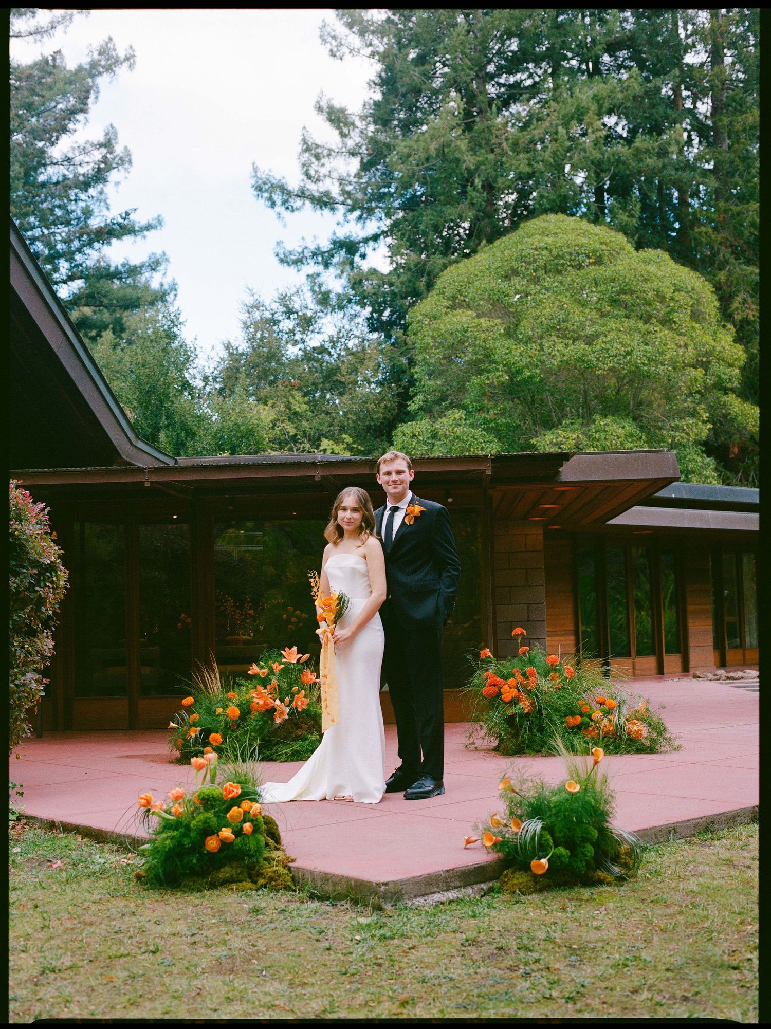 A bride and groom pose together on a red patio surrounded by vibrant orange flowers at a woodland wedding venue.