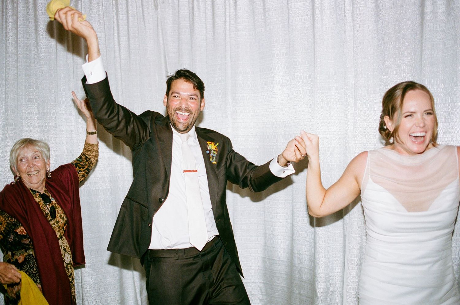 Groom celebrates with arms raised as bride and elderly guest dance at wedding reception.