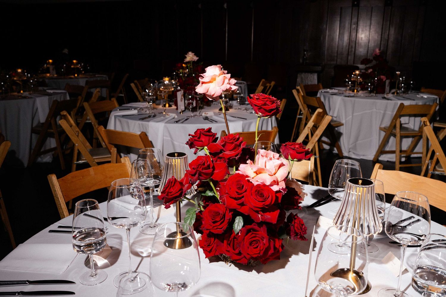 Elegant wedding reception tables with red rose centerpieces, white linens, crystal glassware, and gold accents in a dimly lit venue.