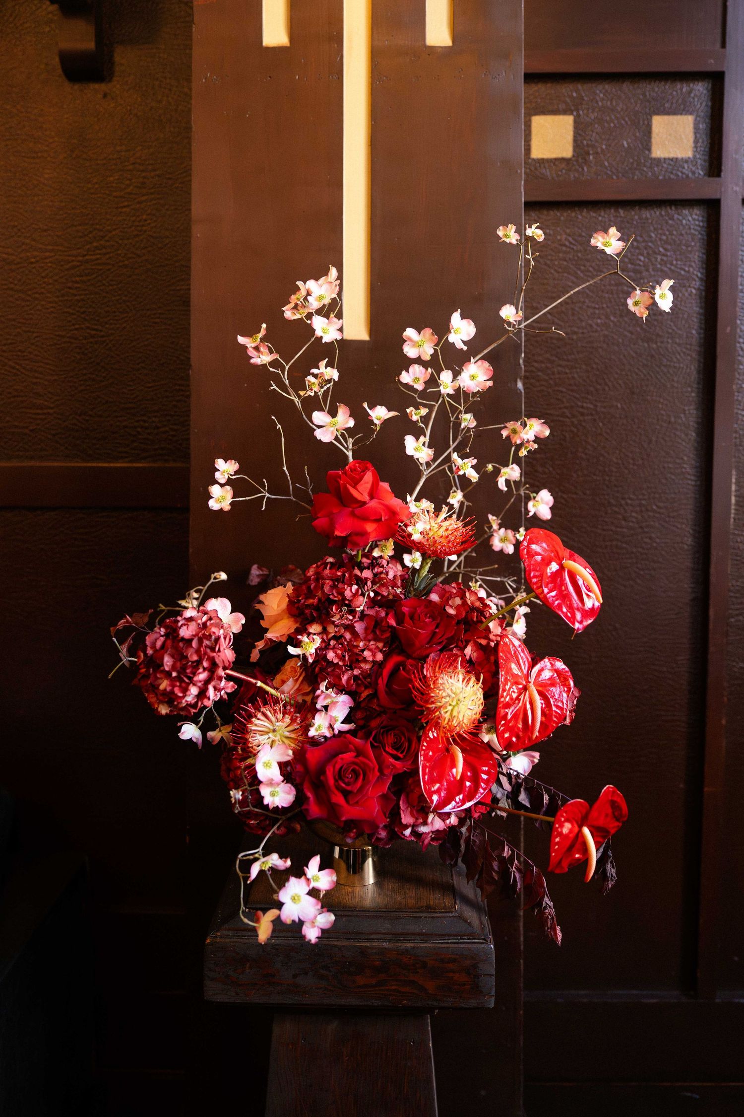 Lush red floral arrangement with roses, anthuriums, and cherry blossoms on a wooden stand against a dark backdrop.