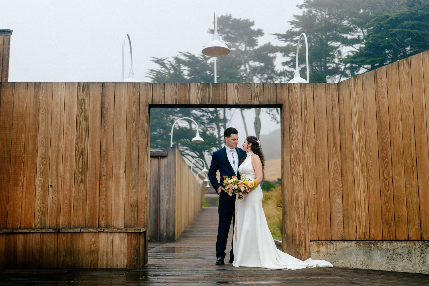 Bride and groom share a romantic moment in a wooden archway on a misty, rainy day at an outdoor wedding venue.
