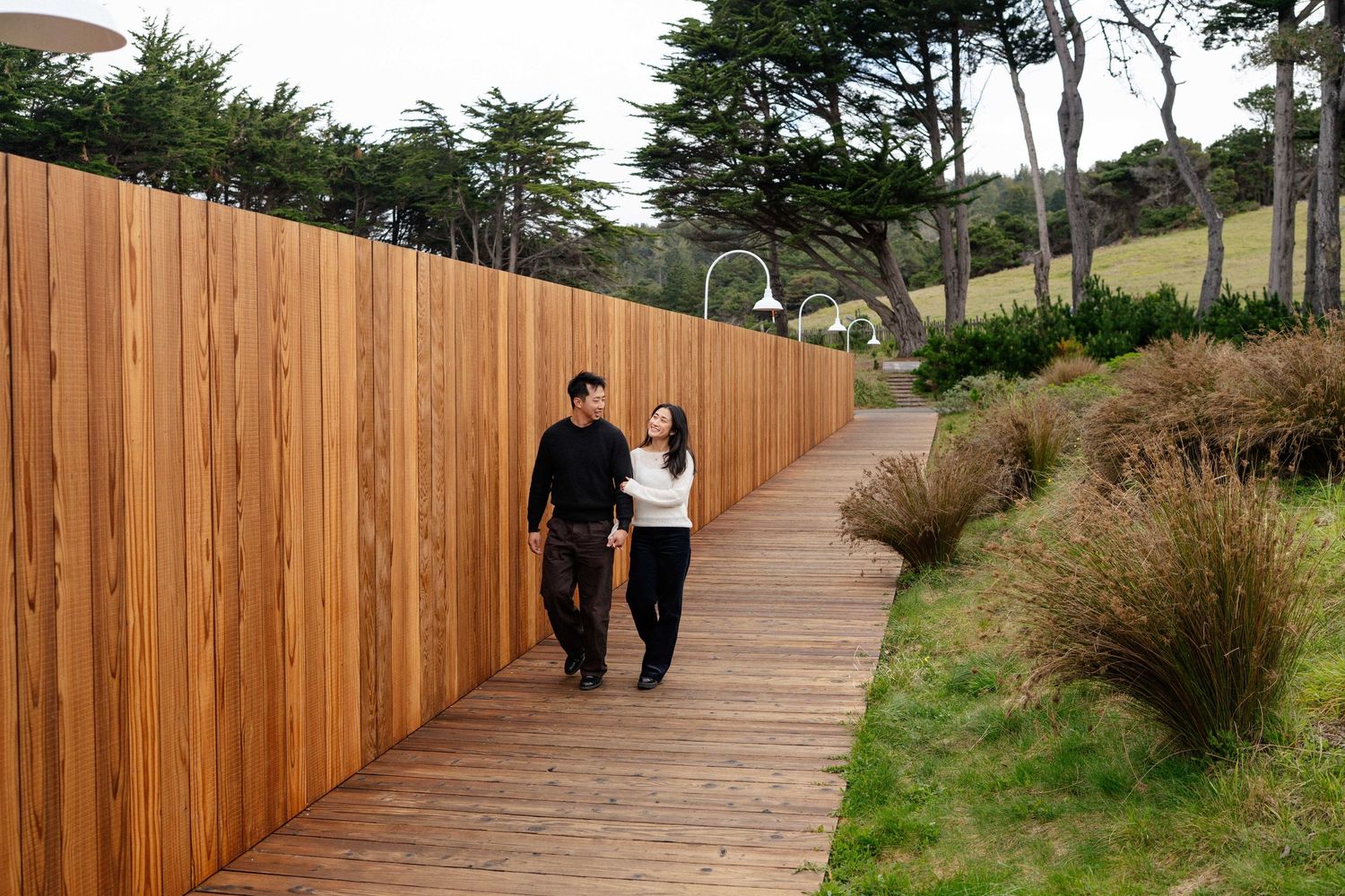 Couple walking hand in hand on a wooden boardwalk path beside a tall cedar fence surrounded by lush greenery.