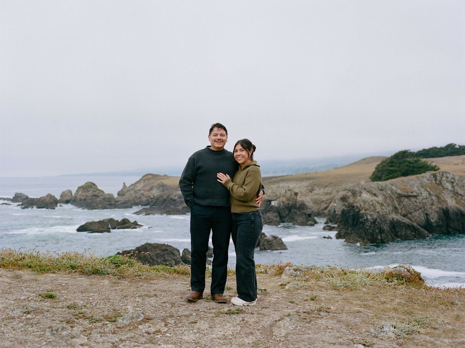 Couple posing together on rocky coastal bluff with ocean and sea stacks in misty background.