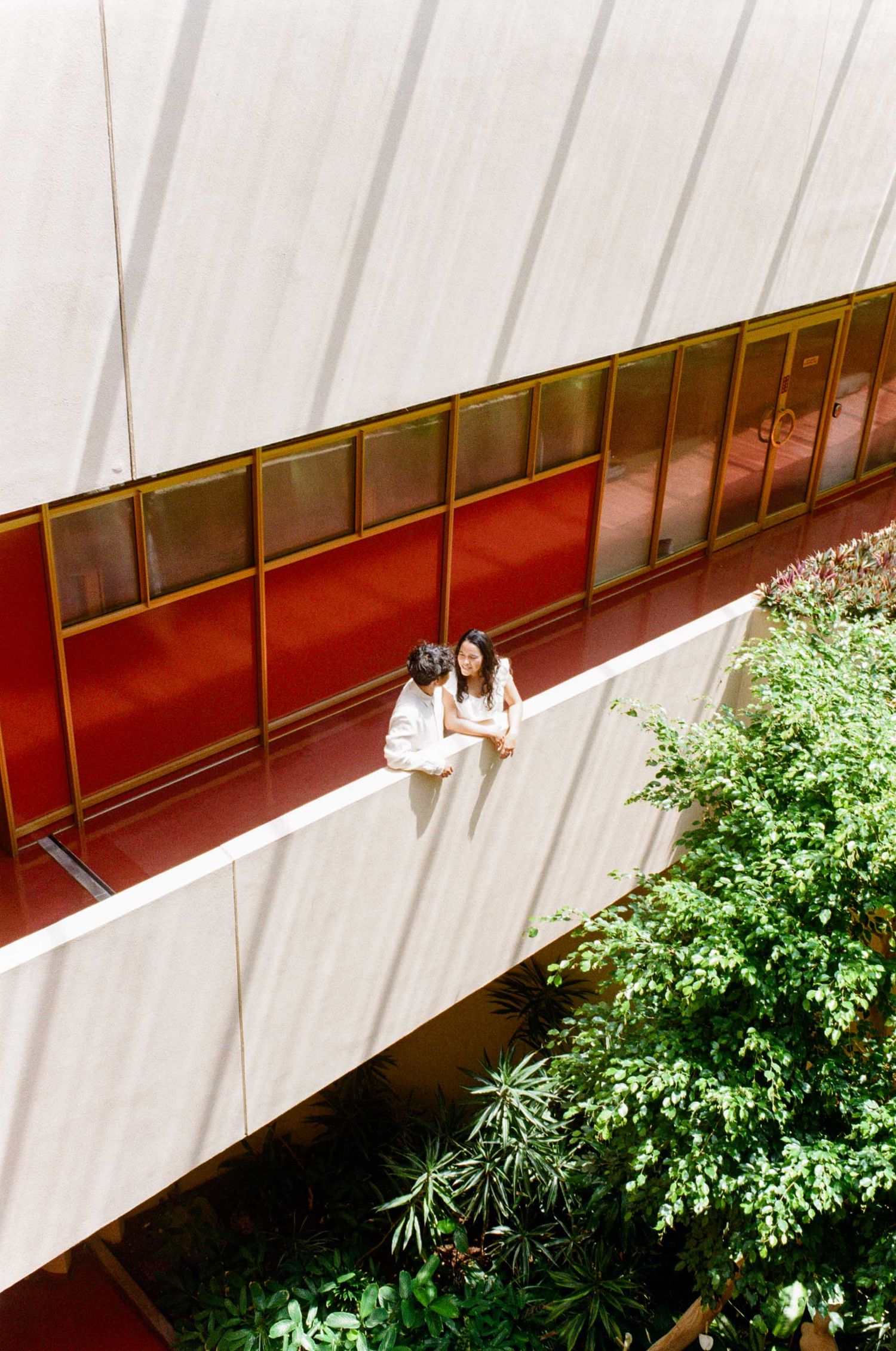 Two women in white dresses stand on a modern building ledge with red panels and lush green trees below.