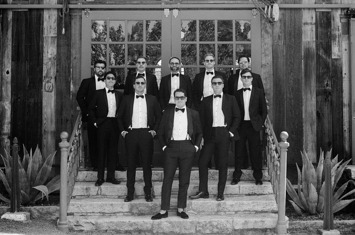 Groomsmen in black tuxedos and sunglasses pose on steps in a black and white wedding photo.