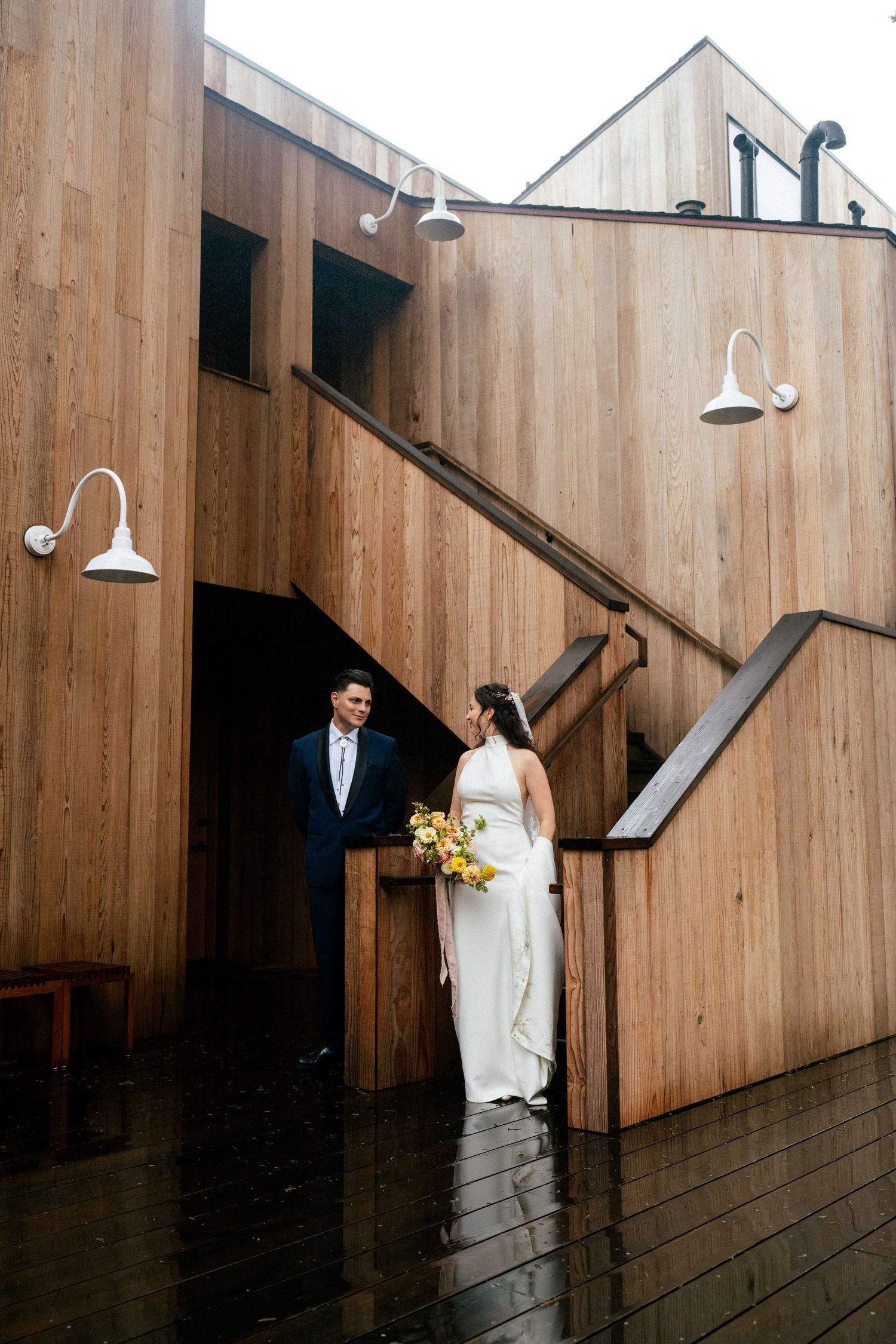A bride and groom share a romantic moment outside a modern wood-clad building on a rainy day.