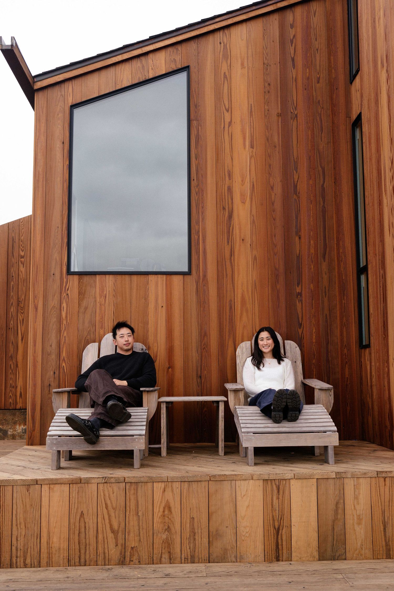 Two people relaxing in Adirondack chairs on a wooden deck outside a modern cedar-clad building.