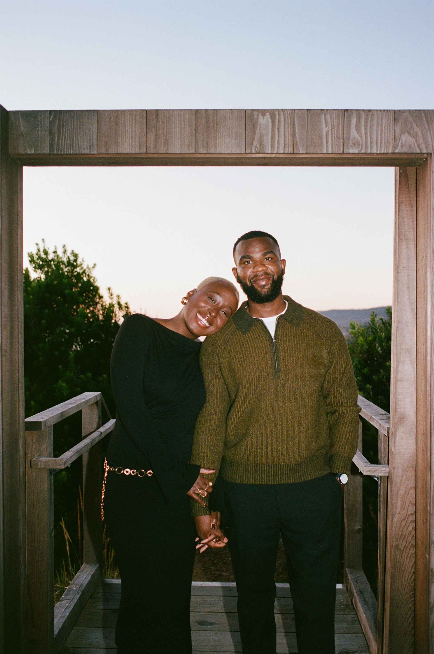 A couple smiles and holds hands on a wooden deck at sunset, framed by a rustic doorway with greenery behind them.