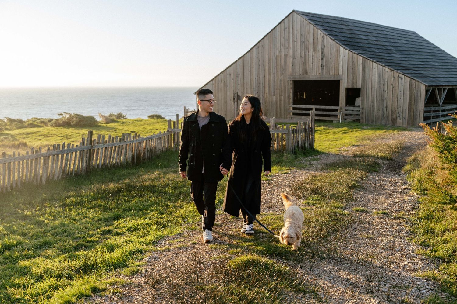Couple walking dog on rural coastal path beside wooden barn at sunset.