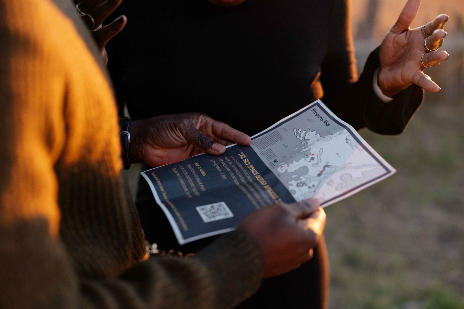 Two people examining a booklet outdoors in warm evening light, one gesturing expressively.