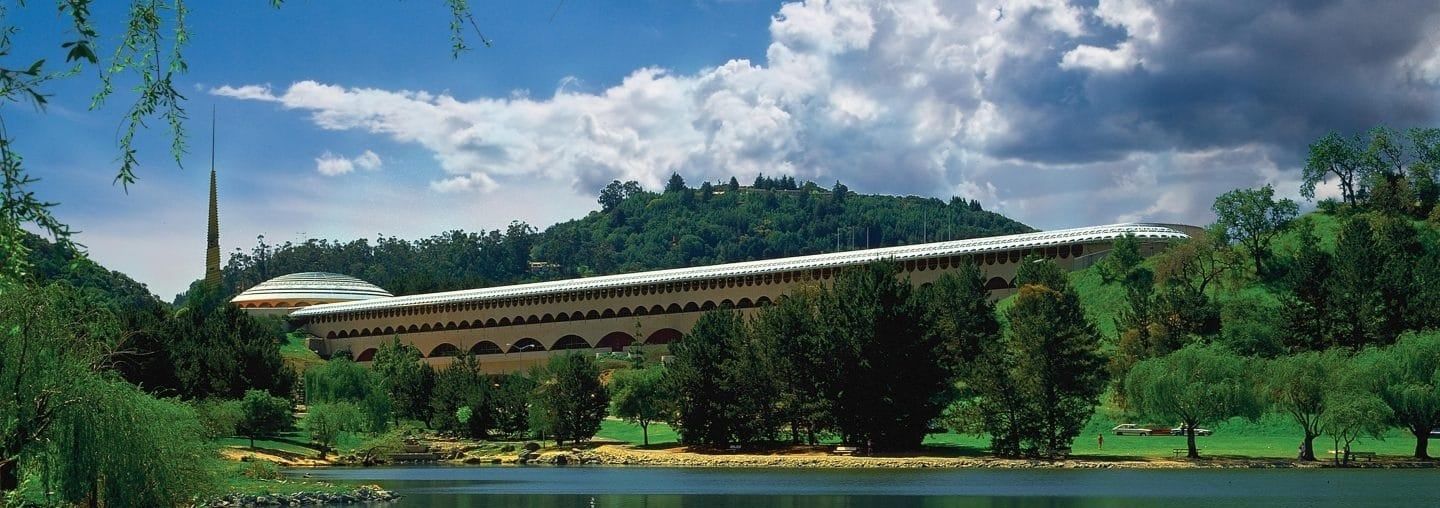 Panoramic view of a modern arched building surrounded by lush green trees and a serene pond.
