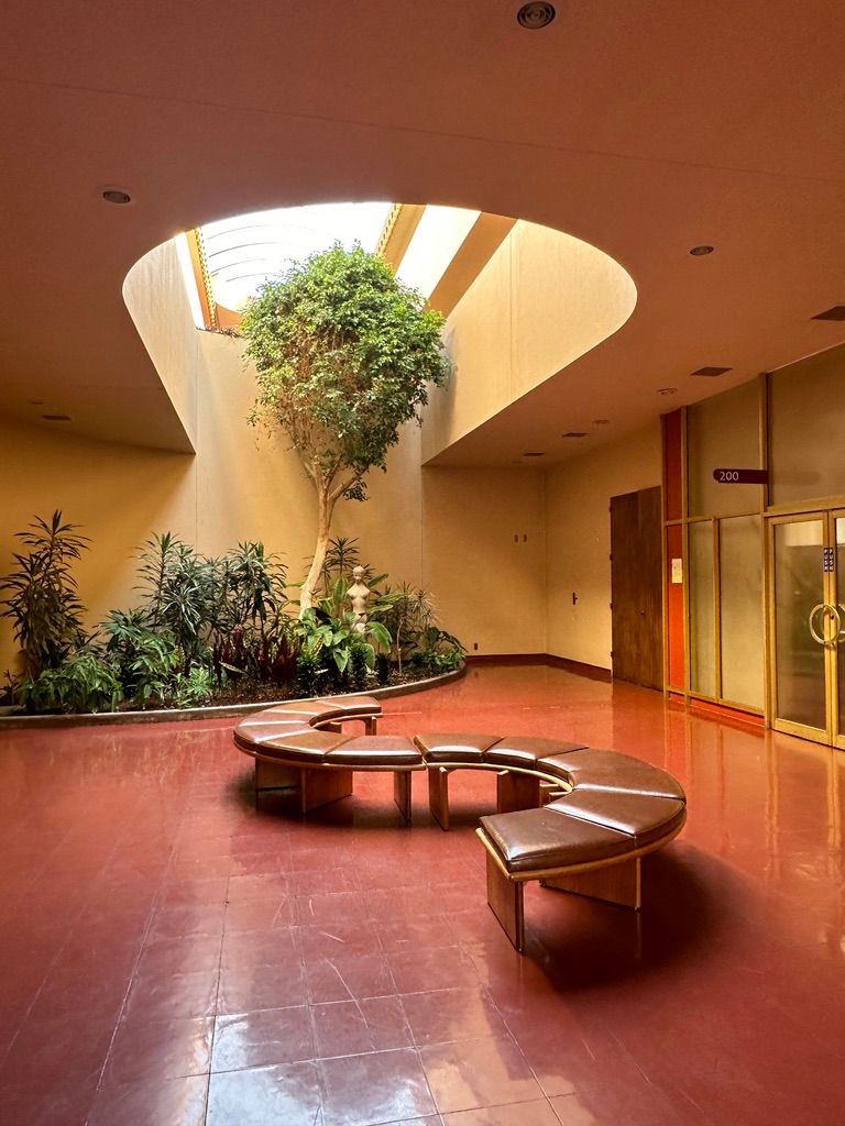 Bright atrium interior with spiral skylight, lush indoor tree, curved benches, and terracotta tile floors.