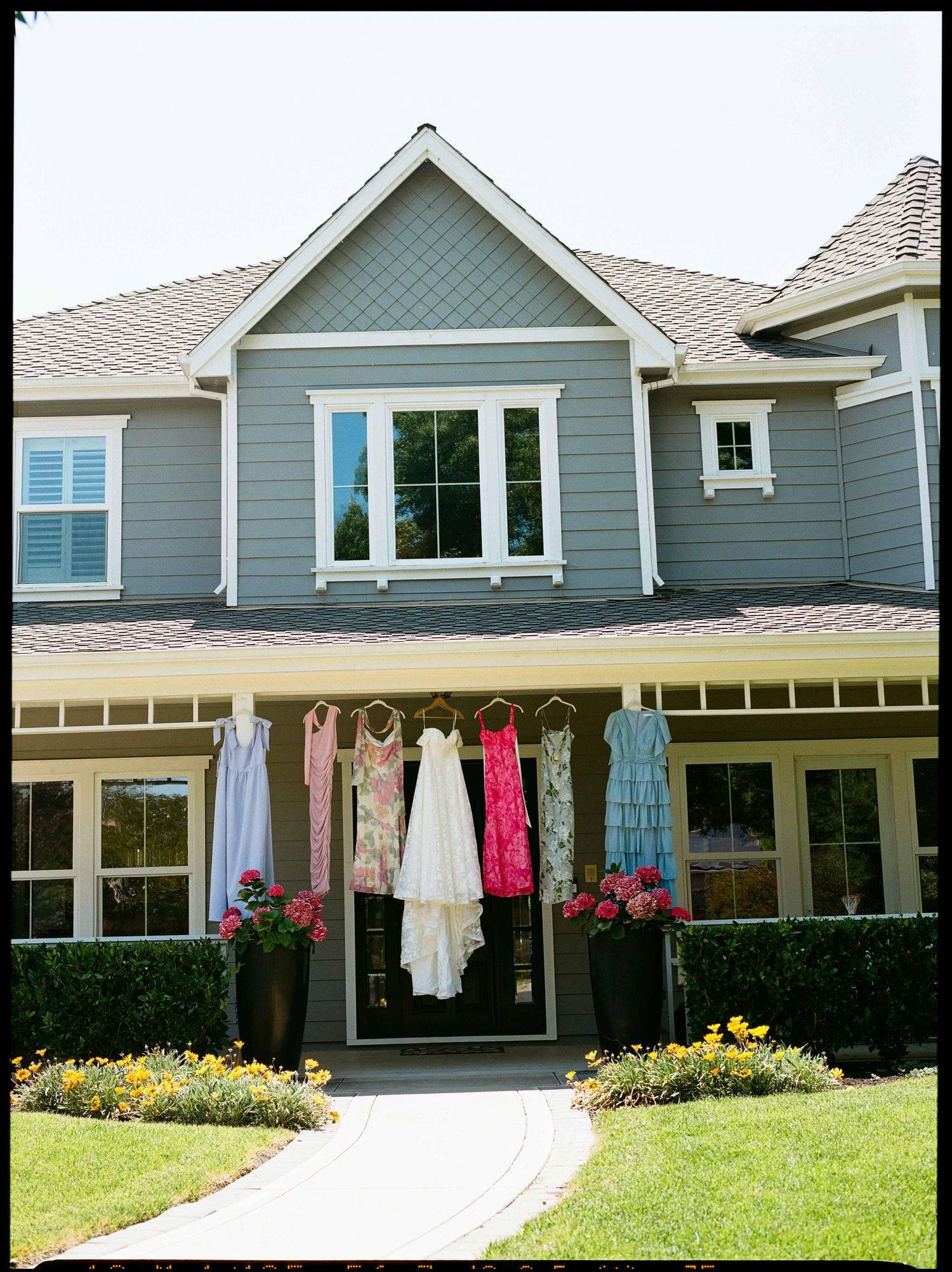 Wedding and bridesmaid dresses hang on gray house exterior for outdoor bridal display.