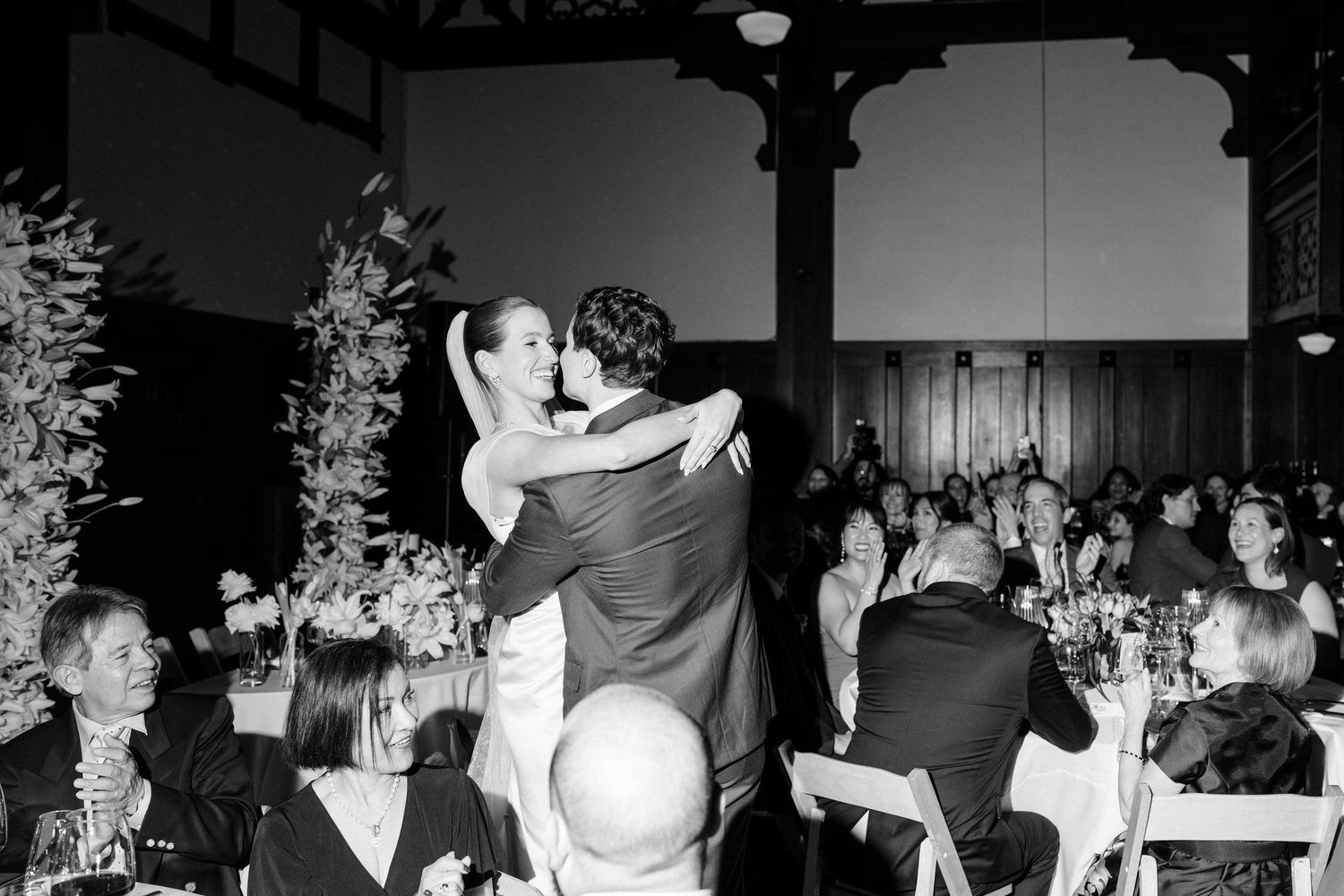 Black and white photo of a couple sharing their first dance at a wedding reception with guests seated at tables.
