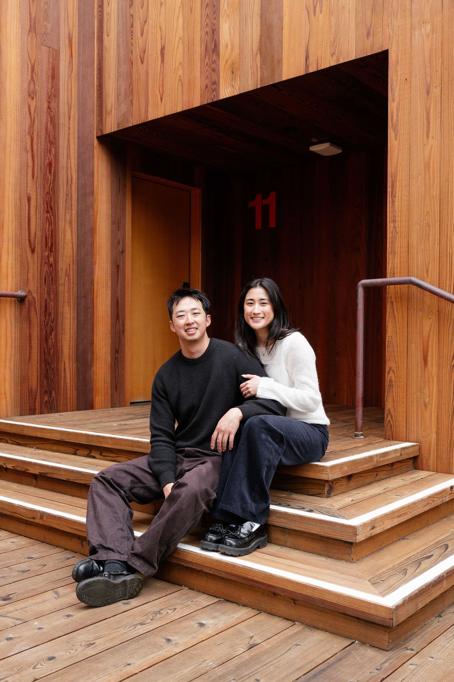 A smiling couple sits on wooden steps outside a modern building with warm cedar wood paneling.