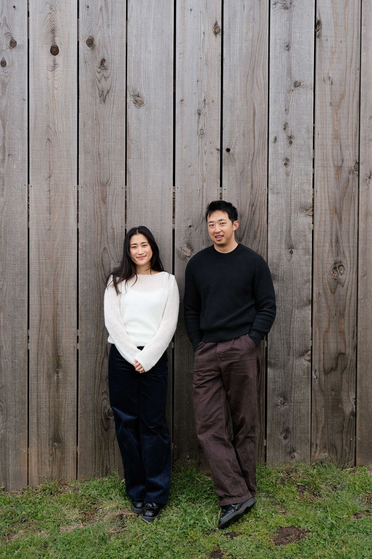 Two people smiling in casual attire stand against a rustic wooden fence on green grass.