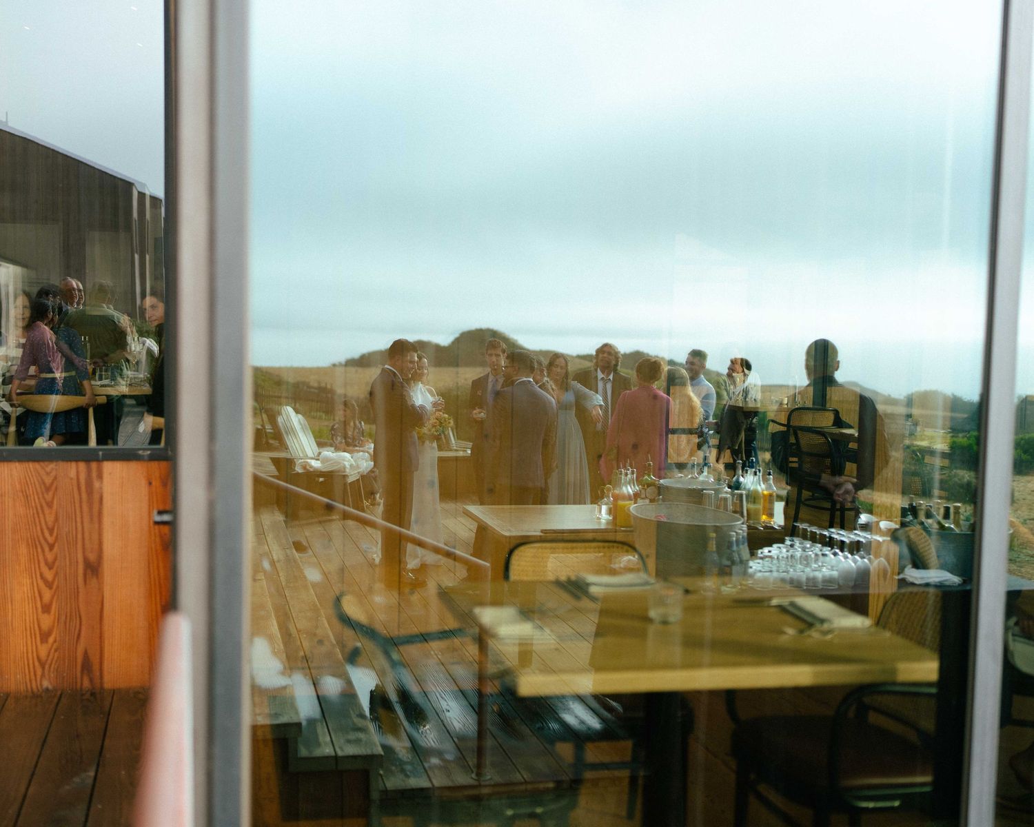 Reflection in glass window of people gathered on outdoor deck with wooden furniture and scenic landscape views.