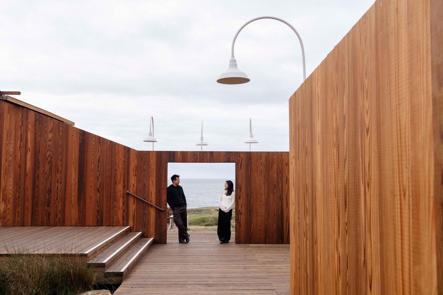 Two people conversing in a stunning timber-clad coastal venue with ocean and sailboat views under an overcast sky.
